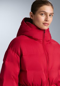 Piros steppelt dzseki magas nyakú kapucnival, sima textúrával és vízszintes steppelt mintázattal, cipzáras rögzítéssel.