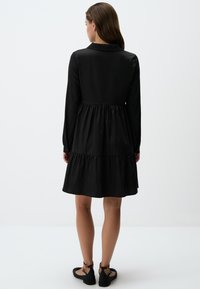 Zwarte lange mouwen jurk met een gelaagde rok, voorzien van een kraag en knoopmanchetten. Glad materiaal met een casual, ontspannen pasvorm. Zwarte platte sandalen.