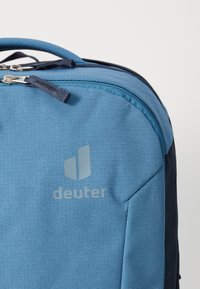 Rucsac din material textil de culoare albastră, cu o finisare texturată, având un panou lateral albastru închis, fermoare argintii și un logo gri proeminent pe față.
