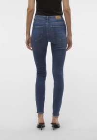 Uske traperice u plavoj boji s glatkom teksturom. Imaju struk srednje visine, suptilno šivanje i stražnje džepove. Nose se s crnim cipelama na petu.