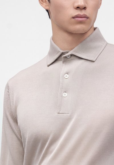 Gros plan d'une personne portant un polo beige à manches longues avec un col plié et deux boutons blancs fermés.