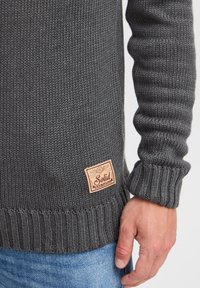 Grå strikket sweater med ribbet halsudskæring og manchetter, med en tekstureret overflade. Inkluderer et rektangulært lædermærke med teksten "Solid."