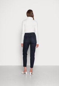 Dunkelblaue Jeans mit schmal zulaufendem Bein, hoher Taille und Detailverarbeitung an den hinteren Taschen. Kombiniert mit einem weißen gerippten Rollkragenpullover und weißen Blockabsatzschuhen.