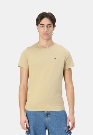 Giovane uomo in piedi, indossa una maglietta beige a maniche corte e pantaloni cargo in denim blu, su uno sfondo chiaro uniforme.