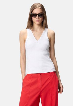 Donna che indossa occhiali da sole a occhi di gatto in tartaruga, top bianco senza maniche con scollo a V e pantaloni rossi a vita alta con le mani nelle tasche.