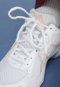 Nærbilde av en hvit atletisk sko med mesh-struktur og snøring, som hviler på en blå overflate. Subtile ferskenfarger er synlige.