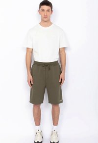 Shorts vert olive avec une taille élastique et un cordon de serrage, assortis à un t-shirt blanc uni. Le mannequin est debout, portant des baskets blanches.