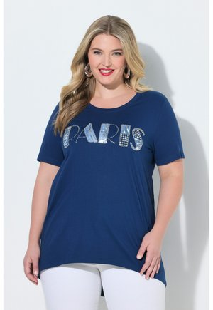 Lachende vrouw met lang blond haar, draagt oorringen, een marineblauw T-shirt met versierde "PARIS"-letters en een witte broek.