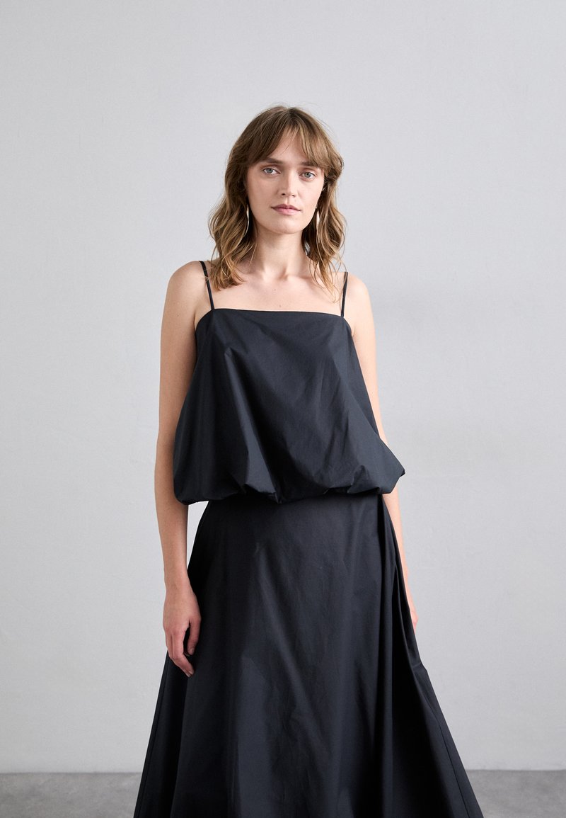 Robe noire avec un corsage drapé et ample, doté de fines bretelles. La jupe est pleine et longue, fabriquée dans un tissu léger avec une texture lisse.