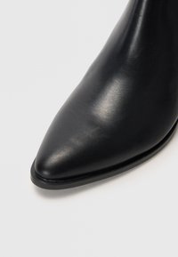 Bota de tornozelo em couro preto com bico afilado e textura suave. Apresenta um brilho subtil e uma sola plana e fina para elevação mínima.