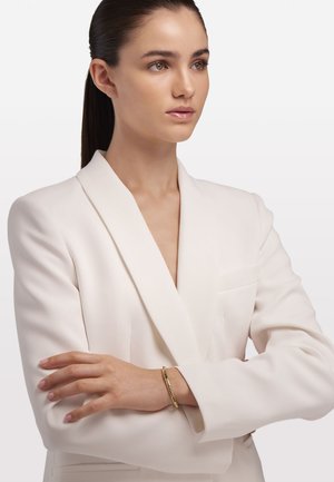 Weißes Blazer mit tiefem V-Ausschnitt, getragen von einem Modell mit verschränkten Armen. Ein dünnes goldenes Armreif wird am Handgelenk getragen. Glatte Stoffstruktur.