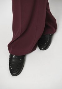 Zwarte instappers met zilveren studs tegen diep mahonie brede broekspijpen. De schoenen hebben een gladde leren afwerking en een afgeronde tekening.