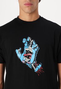 Black cotton t-shirt featuring a graphic design of a blue hand with an open mouth, red tongue, and white teeth. Notable details in print.