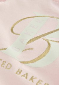 Gaiši rozā audums ar zeltītu izšuvumu un tekstu "BAKED" eleganta fontā. Gluda tekstūra, centrēts dizains un kontrastējošas krāsas.