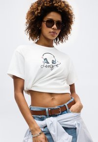 Γυναίκα με γυαλιά ηλίου, λευκό κοντό T-shirt Levi's με γραφικό ακουστικών, μπλε τζιν με καφέ ζώνη και ριγέ πουκάμισο δεμένο γύρω από τη μέση.