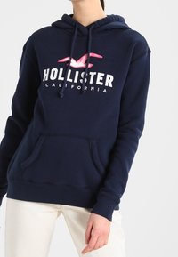Marinblå Hollister-hoodie med rosa måslogo och vit "Hollister California"-text, bärs med ljusbeiga byxor, person beskuren vid nacken.