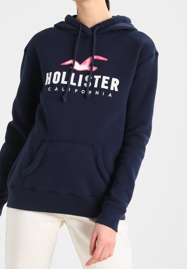 Marinblå Hollister-hoodie med rosa måslogo och vit "Hollister California"-text, bärs med ljusbeiga byxor, person beskuren vid nacken.