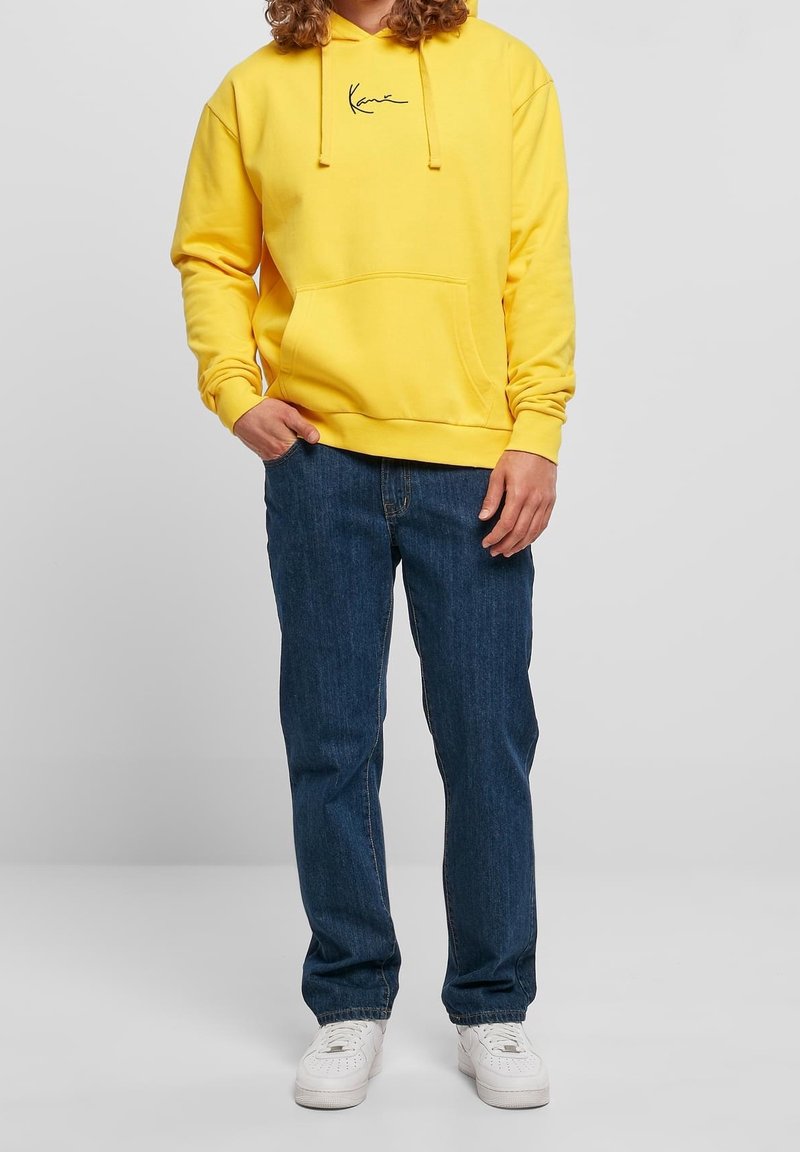 Personne portant un sweat à capuche jaune avec poche avant et logo, jean bleu et baskets blanches, debout devant un fond clair uni.