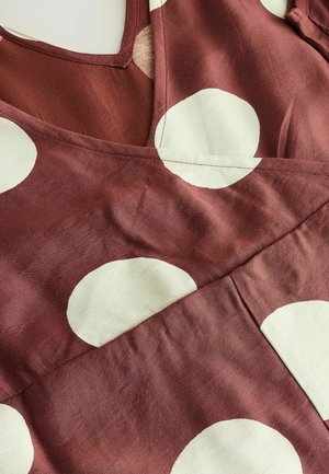 Bordeaux farvet stof med store cremefarvede prikker, udstyret med V-halsudskæring og overlappende lag, der fremhæver en glat tekstur og diagonale syninger.