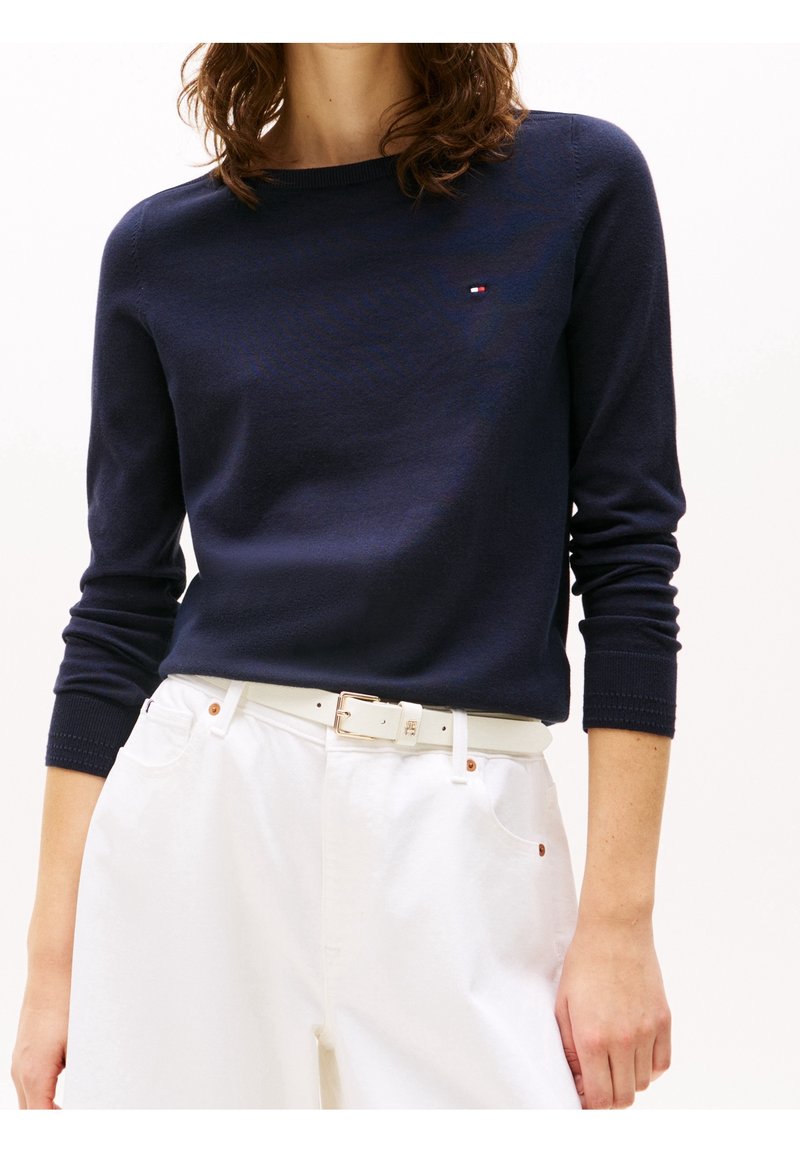 Femme portant un pull bleu marine à manches longues et un pantalon blanc taille haute avec une ceinture blanche, debout devant un fond uni.