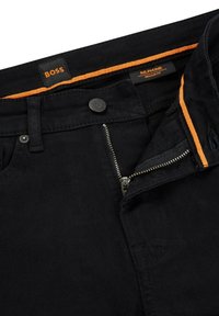 Jean noir avec fermeture éclair partiellement ouverte, taille boutonnée et bordure intérieure orange affichant les étiquettes "BOSS" et "RE.MAINE Regular Fit".