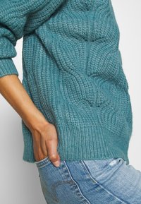 Maglione lavorato a maglia turchese con un motivo a trecce, polsini e orlo a costine. Abbinato a jeans azzurri chiari con un piccolo strappo alla tasca.