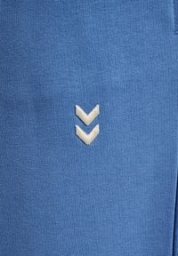 Blå stof med en tekstureret vævning, der har et hvidt broderet chevron-logo nær bunden. Simpelt design, ingen yderligere mønstre.