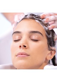 Session de shampooing mettant en scène une personne aux cheveux foncés et ondulés, les yeux fermés, avec une mousse vive et des mains massant doucement le cuir chevelu.