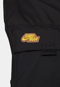 Černý textilní úsek bundy s vyšívaným logem "JUMP MAN" ve žluté a hnědé barvě s texturovaným povrchem a pečlivým prošitím.