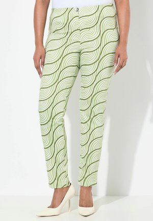 Pantalons verts et blancs à motifs avec un design ondulé, fabriqués dans un tissu lisse, associés à des chaussures à talons hauts crème et un haut blanc.