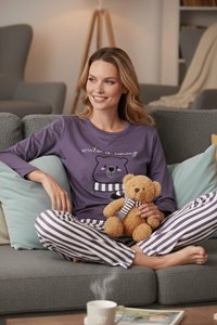 Femme en pyjama violet à motif d’ours tenant un ours en peluche, assise en tailleur sur un canapé gris avec des coussins, souriante et regardant sur le côté.