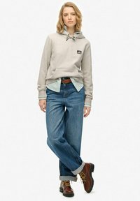 Felpa grigia chiara con cappuccio e tasca frontale, indossata sopra una camicia a righe azzurre. Jeans blu sbiaditi con risvolti e stivali marroni.