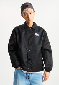 Black, lightweight jacket with snap buttons, relaxed fit, elastic cuffs, and a Vans logo patch. Worn over a white shirt and blue jeans.