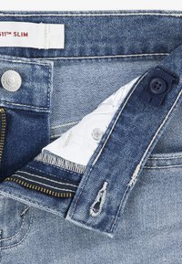 Κολλητό τζιν παντελόνι από μπλε denim με φερμουάρ στο μπροστινό μέρος, μεταλλικό κουμπί και αντίθετες λευκές εσωτερικές τσέπες. Διαθέτει ελαφρύ φθαρμένο εφέ και εμφανείς ραφές.