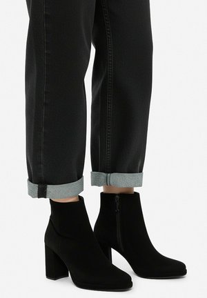 Benen met zwarte omslagjeans en zwarte suède enkellaarzen met blokhakken en zijritsen op een effen witte achtergrond.