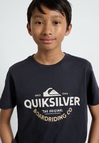 Mørk marineblå t-skjorte med kort erme laget av bomull, med et fremtredende grafisk design i hvit og beige som har teksten "Quiksilver" og et fjellikon.