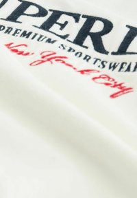 Bijela pamučna majica s izvezenim plavim "SUPERDRY" logotipom i crvenim natpisom "New York City", s mekim teksturom i ležernim dizajnom.