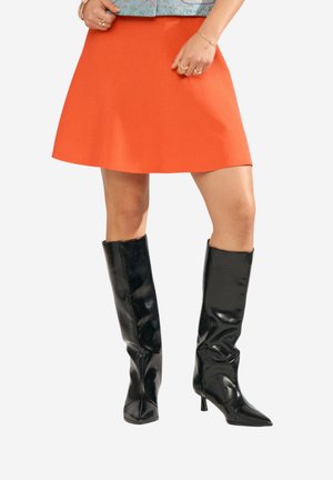 Donna che indossa una gonna arancione a trapezio con stivali neri alti fino al ginocchio, punta a punta e tacco basso, mani sui fianchi, sfondo bianco.