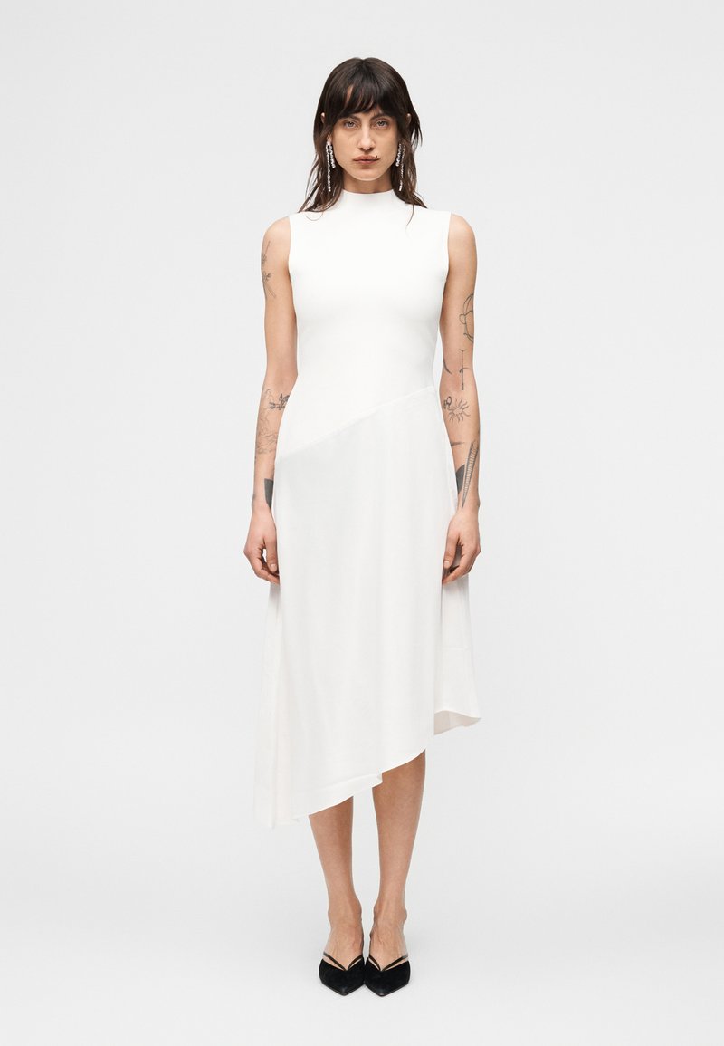 Γυναίκα με τατουάζ που φοράει λευκό αμάνικο ασύμμετρο μεσαίο φόρεμα και μαύρα μυτερά παπούτσια, στέκεται μπροστά σε απλό φωτεινό φόντο.