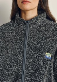 Szara bluza polarowa z teksturowanego, puszystego materiału. Posiada przedni suwak, wysoki kołnierz oraz kolorową naszywkę z logo po lewej stronie.