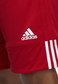 Shorts de sport rouges avec une texture respirante. Présente un logo Adidas blanc et trois rayures blanches sur le côté.