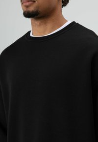 Zwarte sweatshirt met een ronde halslijn en geribbelde afwerking. Heeft een losse pasvorm en zacht katoen, gedragen over een wit T-shirt met ronde hals.