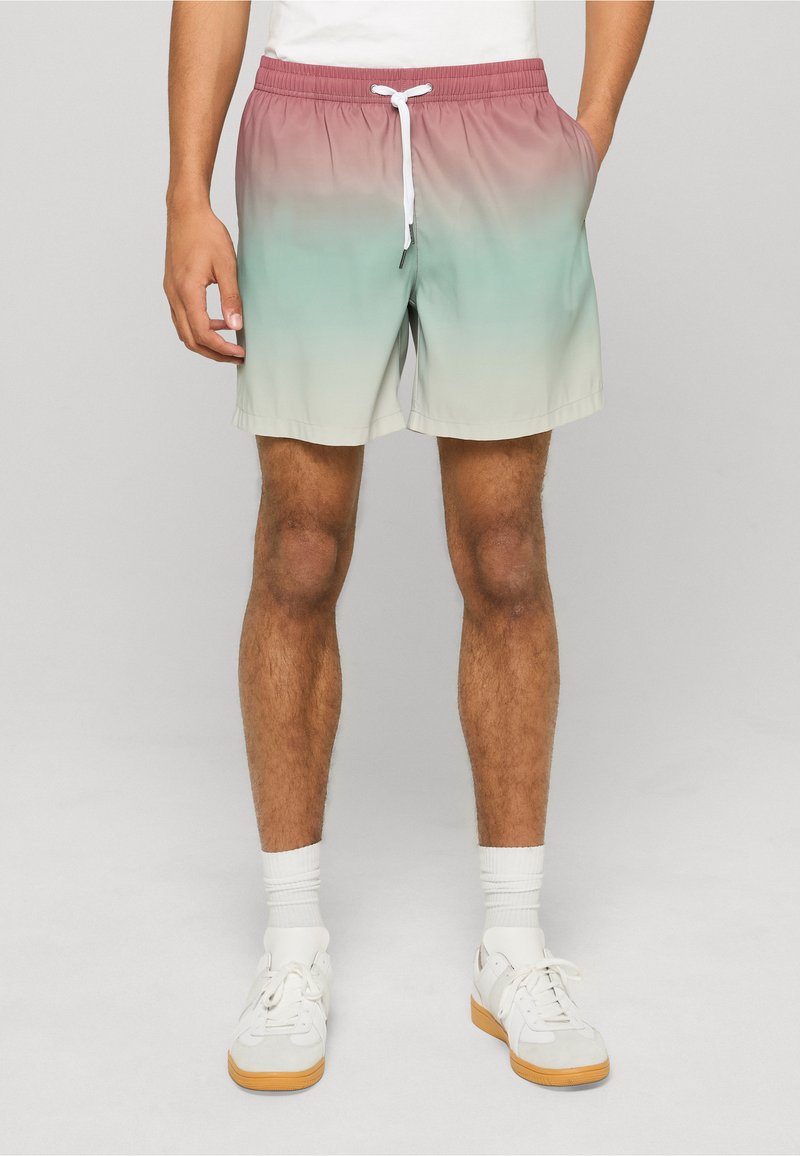 Homme debout portant un short dégradé allant du rose au menthe puis au crème, des chaussettes blanches et des baskets blanches avec des semelles en gomme.