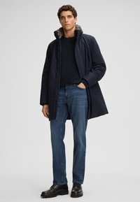 Cappotto navy con colletto alto, foderato internamente in modo morbido, abbinato a jeans blu e scarpe nere stringate, in piedi contro uno sfondo neutro.