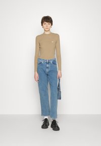 Beige geripptes Langarmoberteil, hoch geschnittene hellblaue Jeans und schwarze Schnürschuhe. Das Modell hält eine kleine Denim-Tasche.