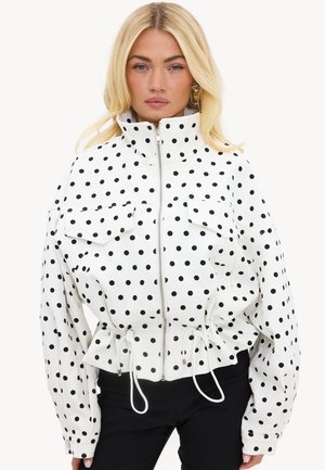 Femme blonde portant une veste blanche à pois noirs, col montant, fermeture éclair à l'avant, taille à cordon et poches à rabat, associée à un pantalon noir.