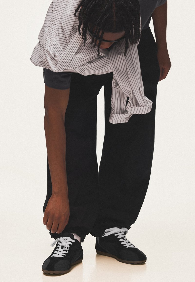 Personne portant un pantalon noir, des baskets noires et une chemise rayée, se penche pour nouer les lacets de sa chaussure gauche.