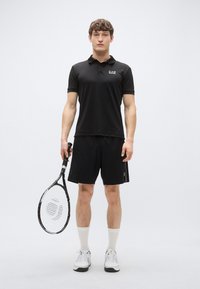 Čierne polo tričko s golierom, v kombinácii s čiernymi krátkymi nohavicami. Model drží tenisovú raketu. Bielé ponožky a športové topánky dopĺňajú outfit.