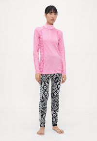 Rosa langermet treningstop med glidelås, med et teksturert mønster. Svart leggings med intrikate hvite blomster- og geometriske mønstre.