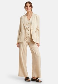 Beige streepjasje met een gestructureerde blazer, knopenvest en wijde broekspijpen. Materiaal lijkt licht en textuur te hebben. Zwarte sandalen.